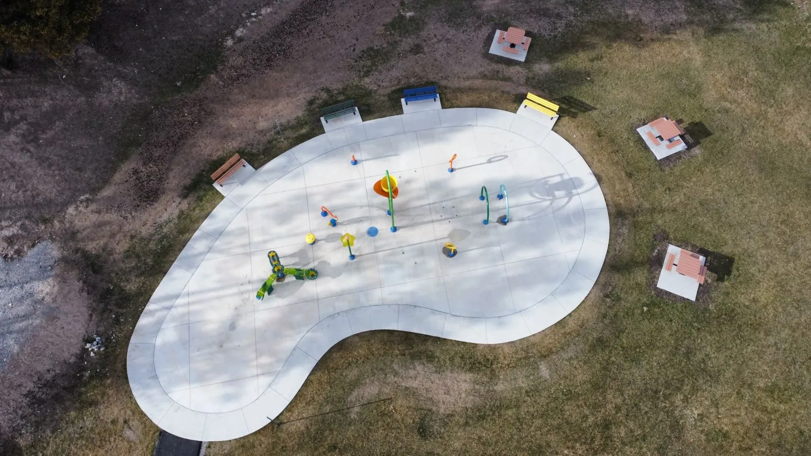 Outdoor benches and picnic tables surround a splash pad. The site furniture provides seating and dining options for visitors to the recreational water play area.