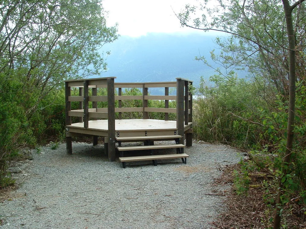 Observation deck constructed with recycled plastic lumber in a natural setting. The deck features a railing and steps, offering a scenic viewpoint surrounded by trees and mountains in the background.