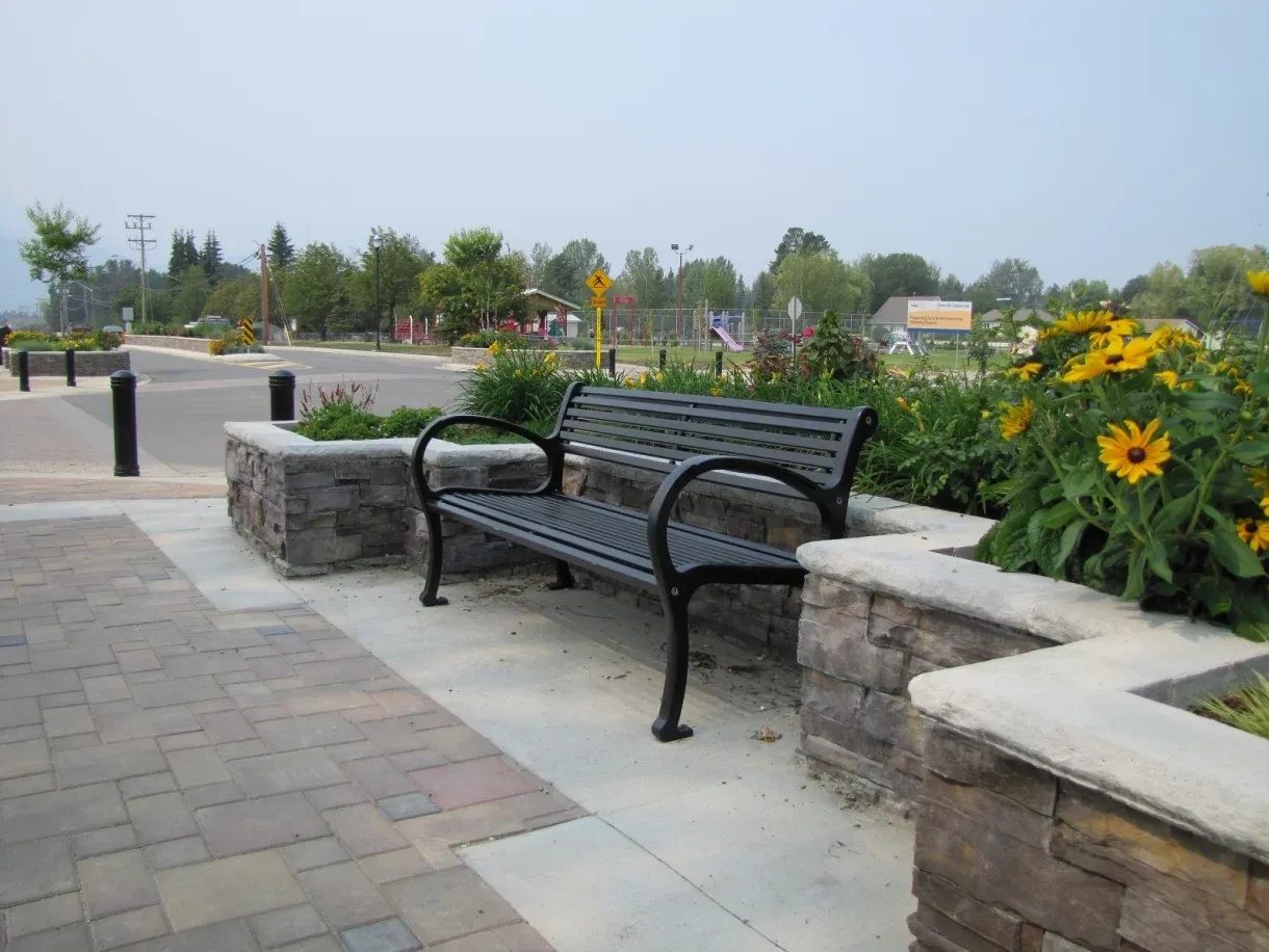 A Mountain Classic bench provides seating along a paved walkway, surrounded by planter boxes filled with flowers and greenery. The bench offers a place to rest near a playground and community space.