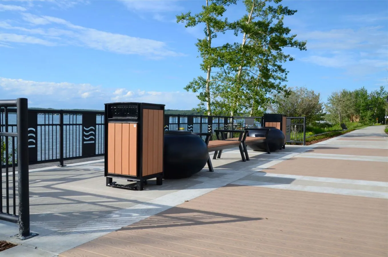 Outdoor setting featuring a Rutherford straight bench and a Beselt square top waste receptacle. The site furniture is installed along a waterfront walkway with decorative railings.
