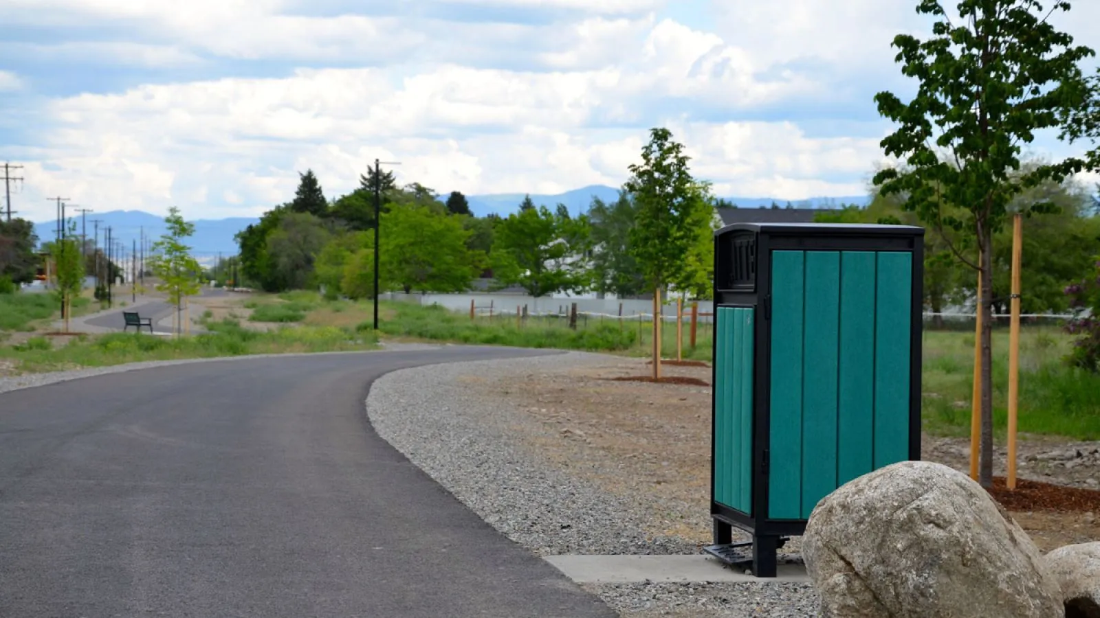 A Beselt square top waste receptacle sits along a paved path in a park-like setting. The bin features green recycled plastic lumber and a black powder-coated frame.