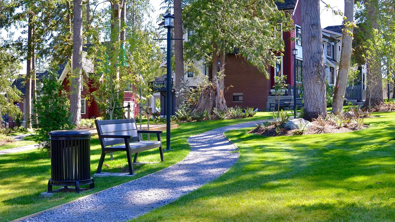 The Beselt round waste receptacle and Rutherford bench sit along a gravel path in a park-like setting. The bench features recycled plastic lumber.