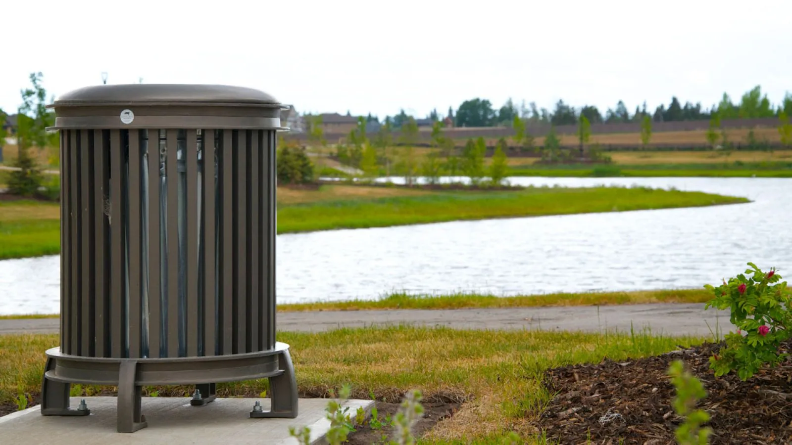 Beselt round waste receptacle in a park setting. The litter bin features a brown powder coat finish and is placed near a paved path and a pond.