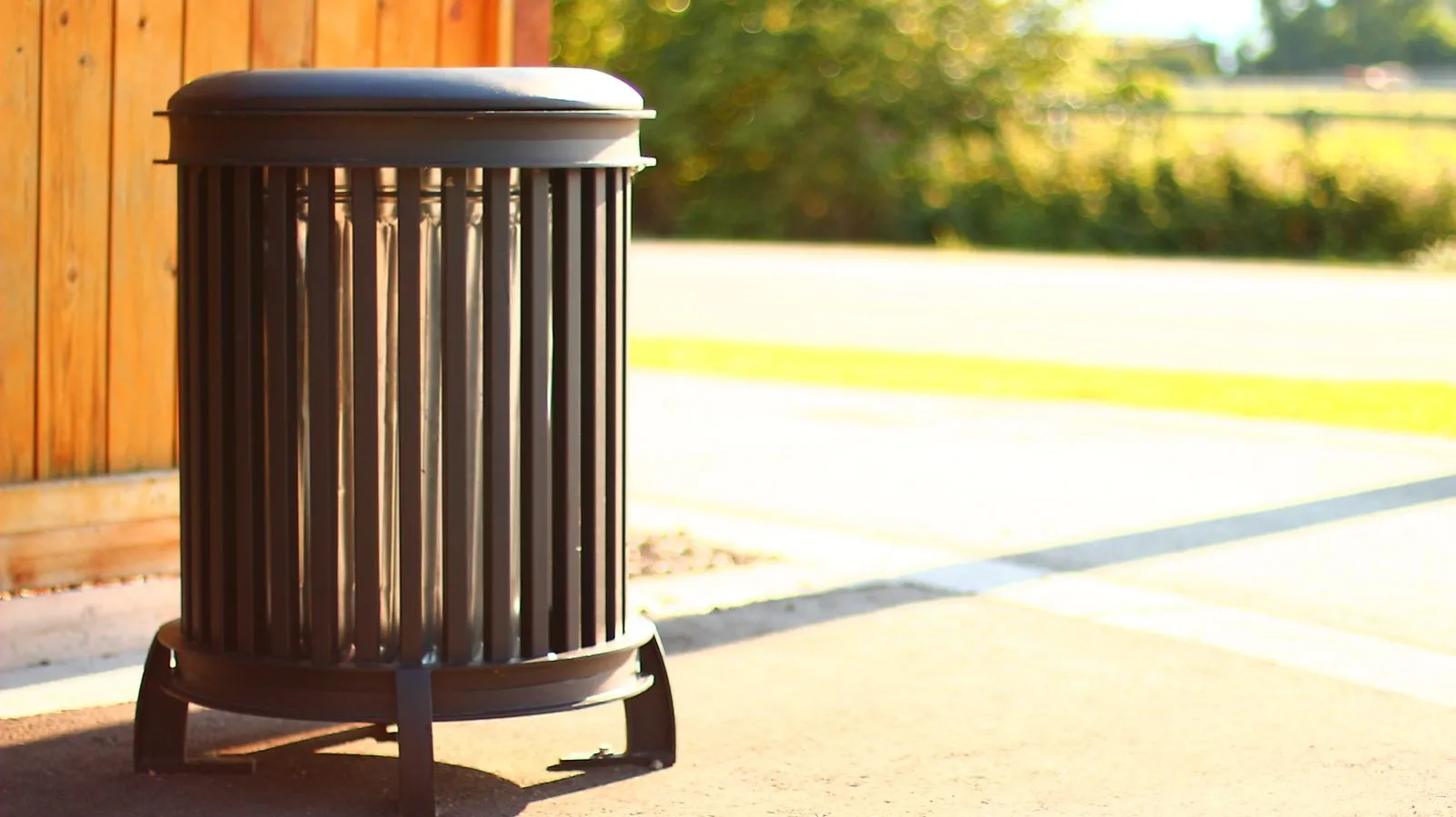 A Beselt Round Waste Receptacle stands on a paved surface near a wooden fence, providing a convenient disposal solution for public spaces.