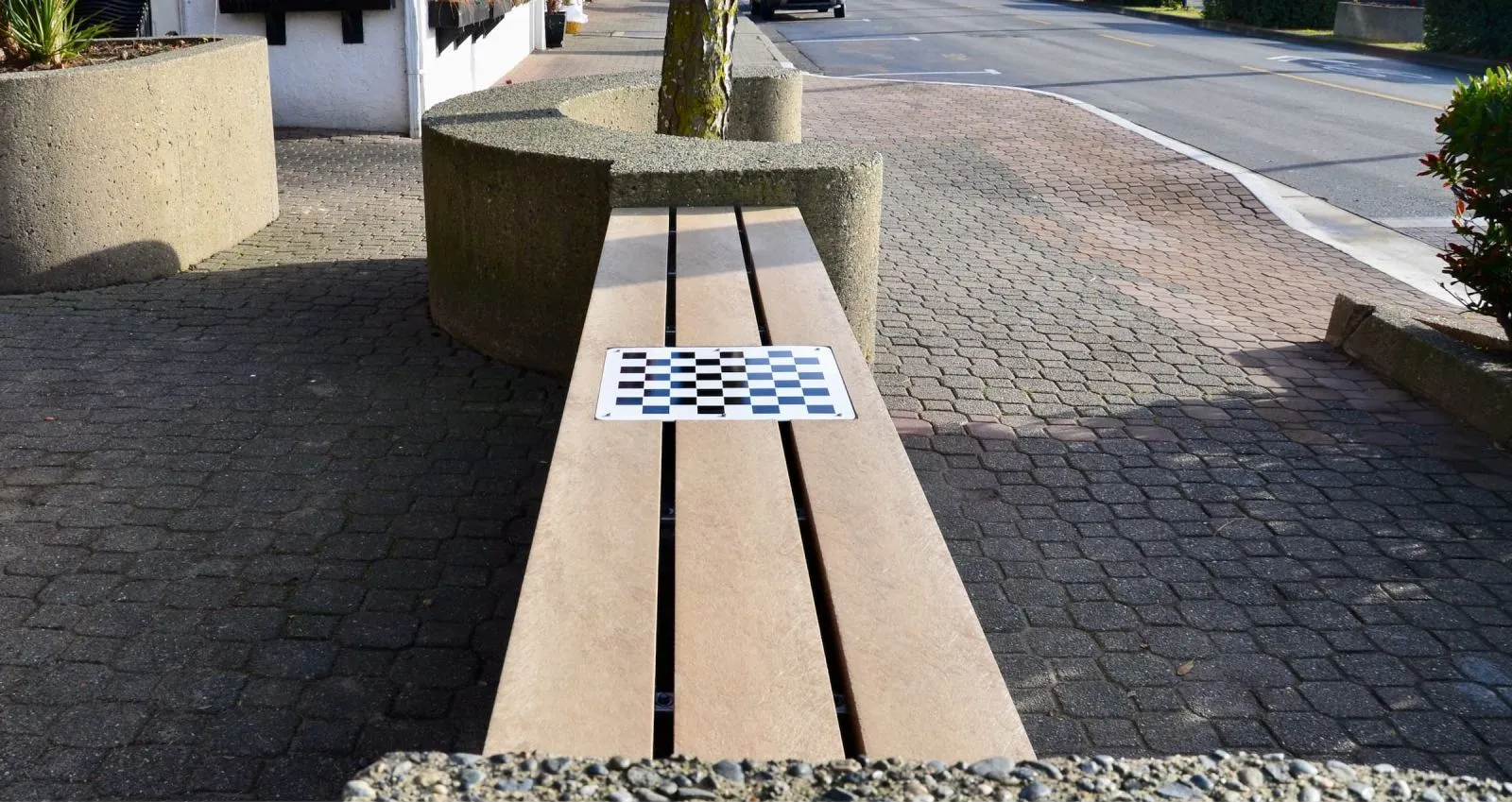 A backless bench with a chess board sits along a paved walkway. The bench features recycled plastic lumber and is situated near concrete planters and a street.