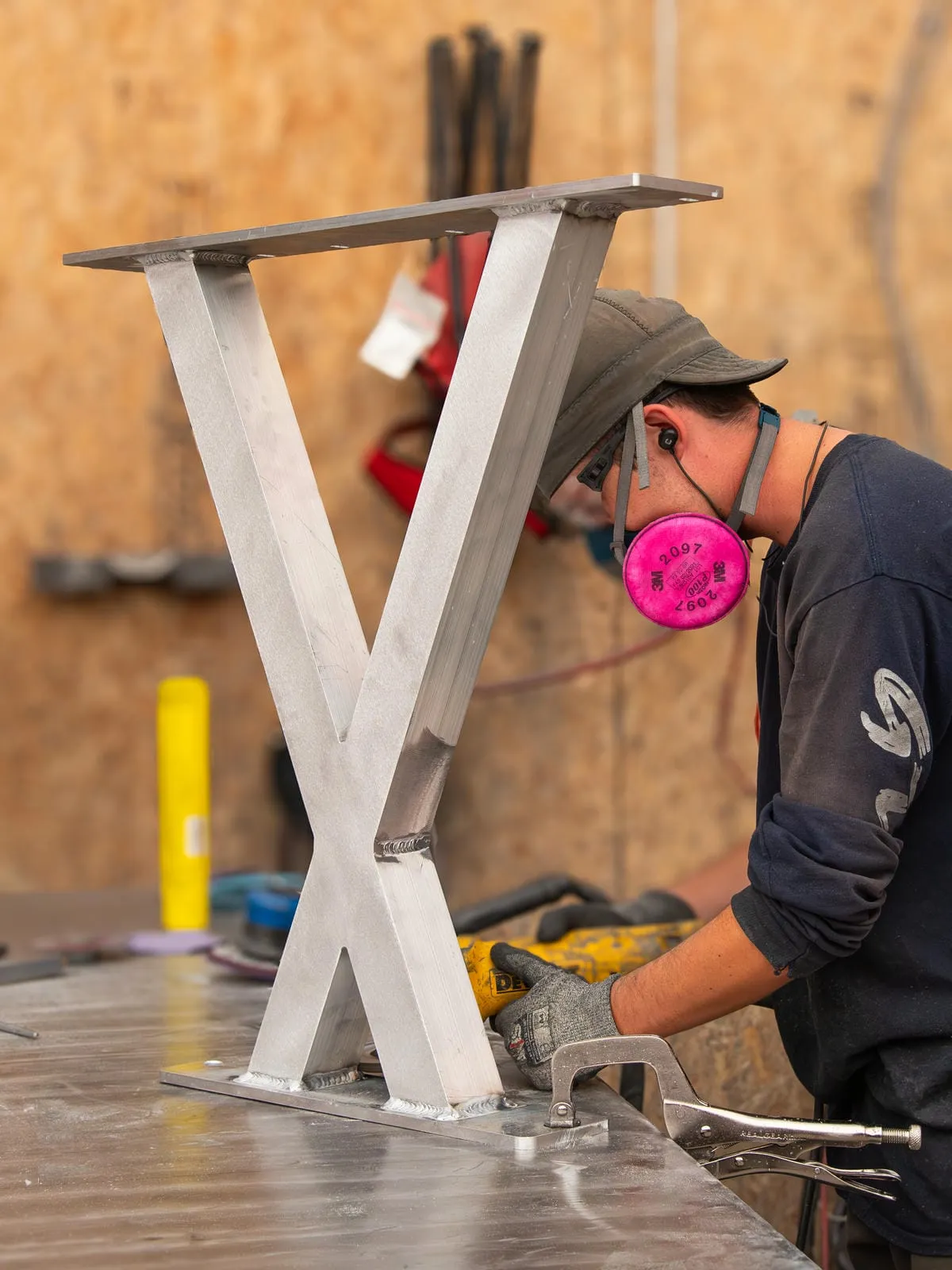 A metal Bayview Picnic Table leg is being cleaned up in a workshop, showing the welding and fabrication process. The leg is silver, and a worker is using a grinder to smooth the welds.