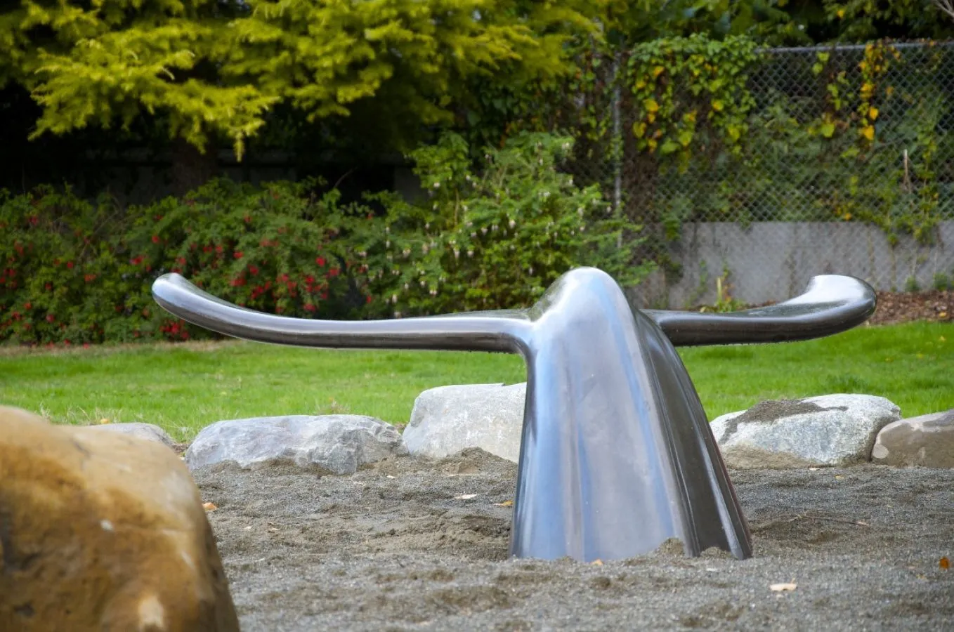 A concrete whale tail bench is partially submerged in a sand pit surrounded by grass and rocks. This unique bench adds an artistic touch to any park or playground.