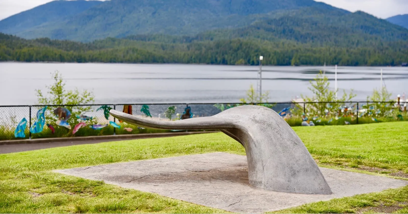 A concrete whale tail sculpture sits on a grassy lawn with a lake and mountains in the background. This unique bench provides a fun and functional seating option for public spaces.