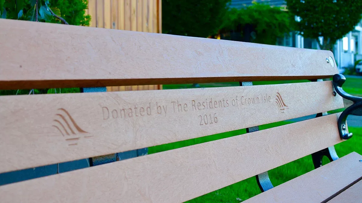 A bench features custom routering with a logo and text indicating it was donated by the residents of Crown Isle in 2016. The bench is made with recycled plastic lumber.