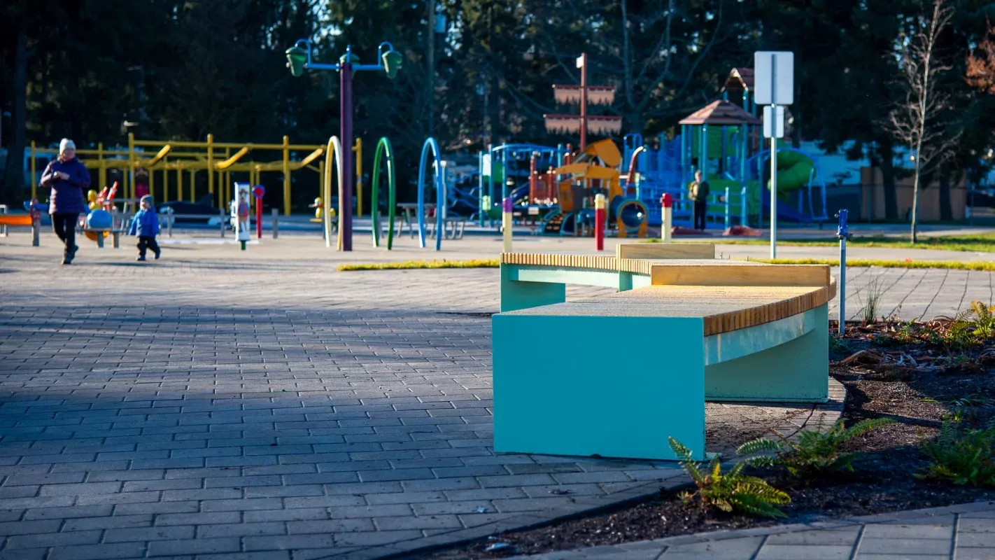 Curved backless bench seating in a park setting. The bench features a light-colored timber seat and a teal-colored base.