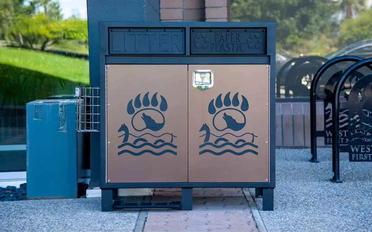 A dual-stream Modena waste and recycling station with custom door panels featuring a wolf and lake design. The unit has a black frame and bronze-colored door panels.