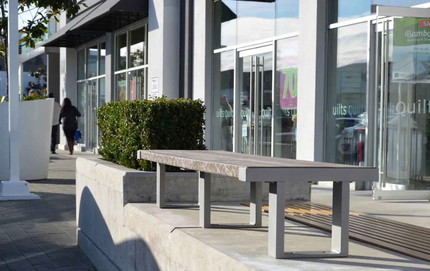 Aylesbury bench with a concrete base sits outside a retail store. The bench features a gray powder-coated frame and recycled plastic lumber.