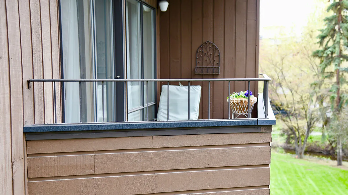 A balcony with brown siding and a black metal railing overlooks a green lawn and trees. The deck is finished with a dark top cap.