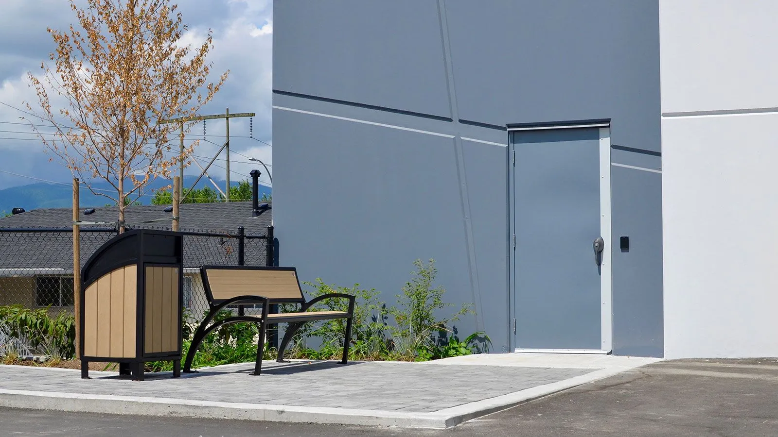 Outdoor setting featuring a bench and waste receptacle. The bench has a black frame and wood-look recycled plastic lumber, while the waste receptacle has a black top and sand-colored sides.