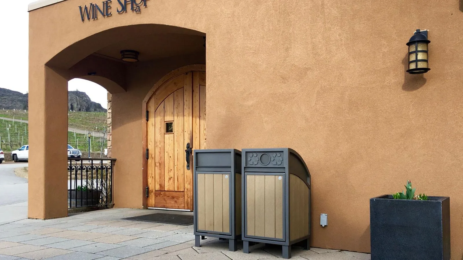 Two Modena Curved Top Waste Receptacles stand outside a wine shop. These bins feature recycled plastic lumber panels and a durable powder-coated metal frame.