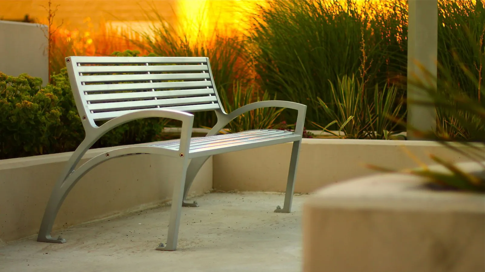 This metal park bench features a sleek, modern design with horizontal slats and curved armrests. It provides comfortable seating in an outdoor setting with lush greenery.