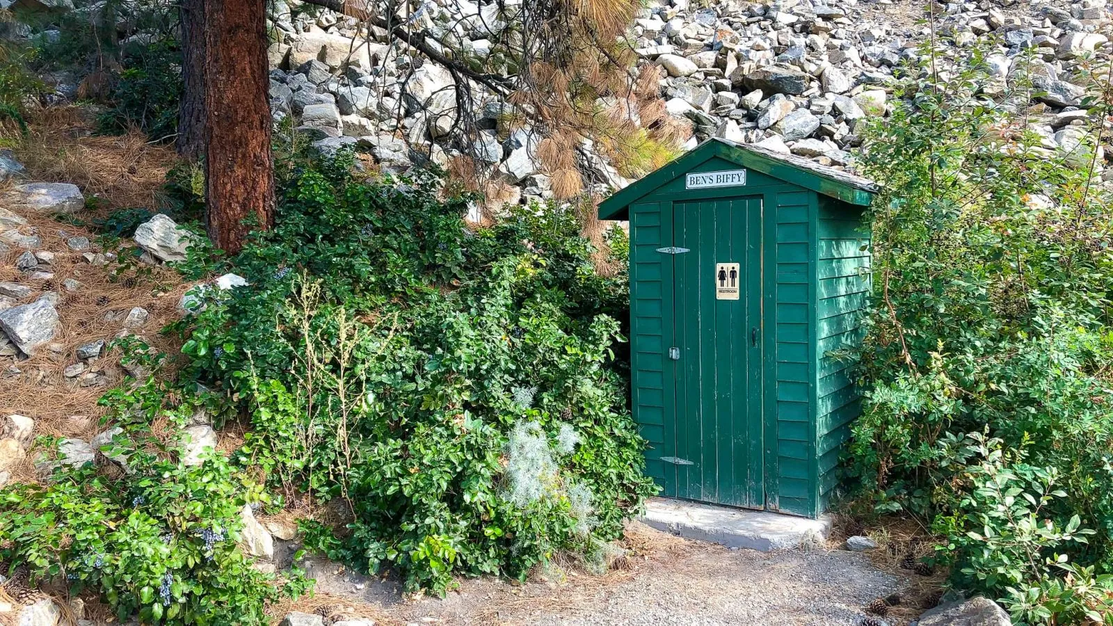 A green outhouse stands nestled among lush greenery and rocky terrain. The structure provides a rustic restroom facility in a natural setting.