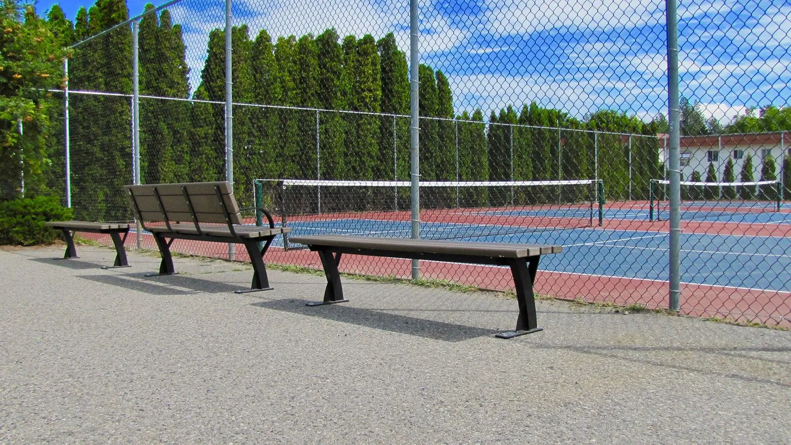 Benches line a walkway next to a tennis court. The benches feature recycled plastic lumber and powder-coated metal frames.