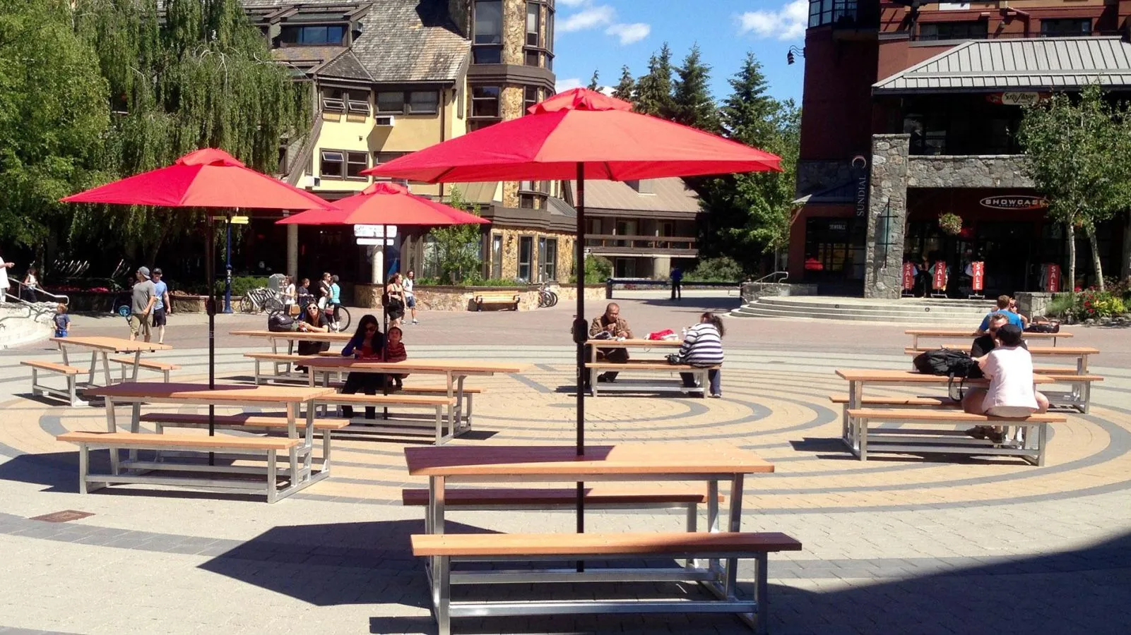 Several Rutherford picnic tables with umbrellas provide outdoor seating in a pedestrian plaza. The tables feature a metal frame and light-colored lumber.