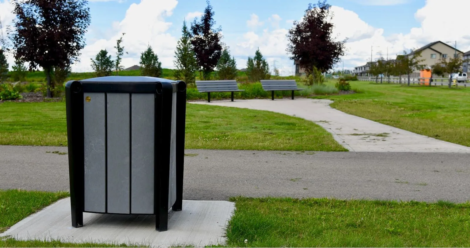 Durable waste receptacle in a park setting with benches and walking paths. The bin features recycled plastic lumber and a powder-coated metal frame.