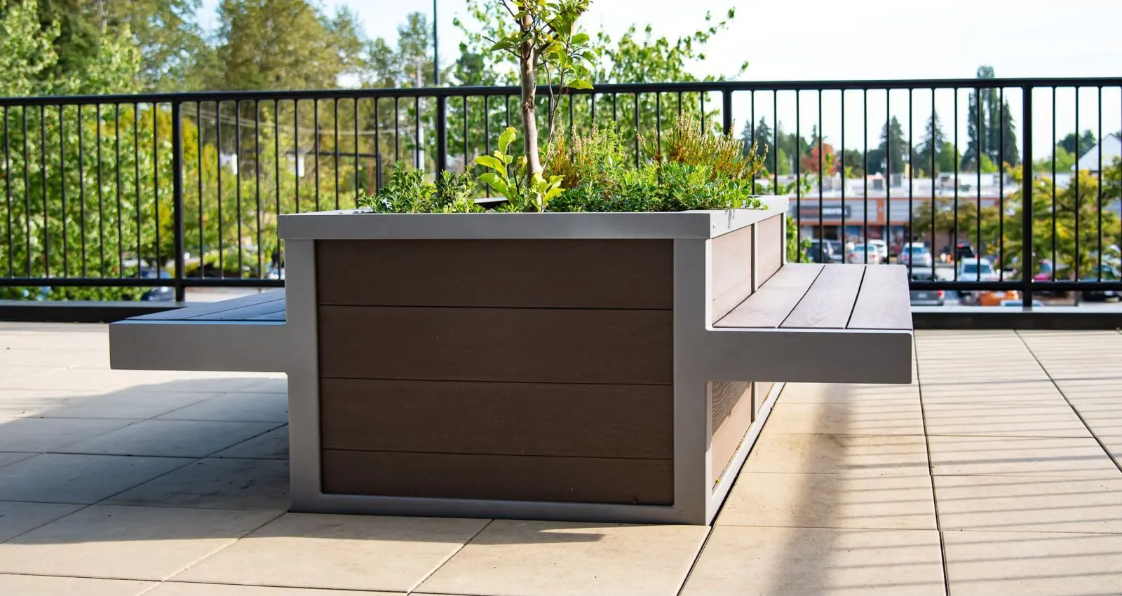 A planter bench combo with seating on either side provides a place to sit and enjoy greenery. The planter box is filled with plants, and the frame is powder-coated in a light gray.