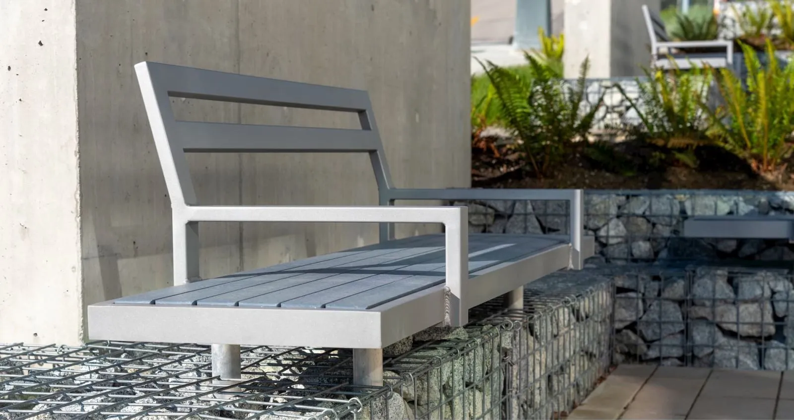 Skyline bench with a light grey powder-coated frame and grey recycled plastic lumber slats. The bench is installed in an outdoor setting with gabion walls and greenery.