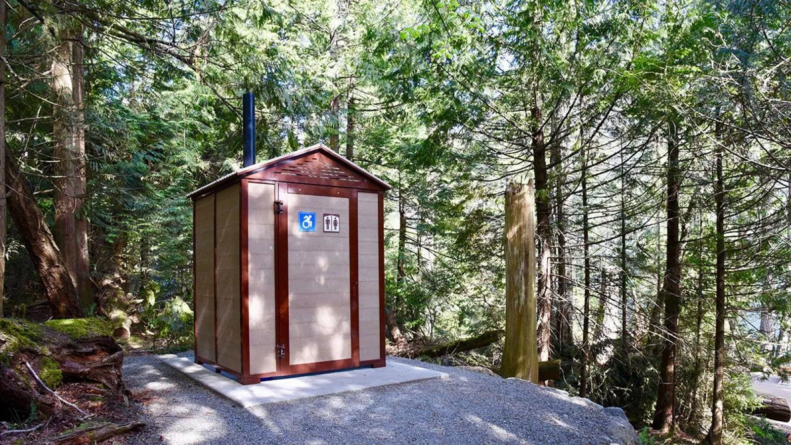 A single-stall outhouse stands in a wooded area. The structure features a light-colored exterior with dark brown trim and a small roof overhang.