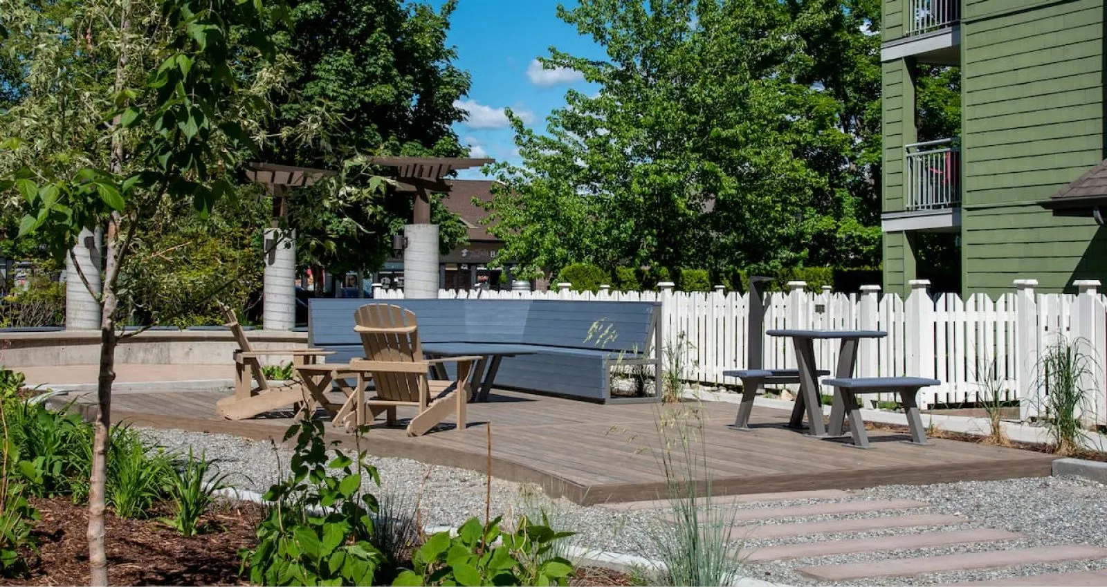 Outdoor seating area featuring a Larson Picnic Table and Adirondack chairs on a wooden deck. The setting includes landscaping, a white picket fence, and a pergola.