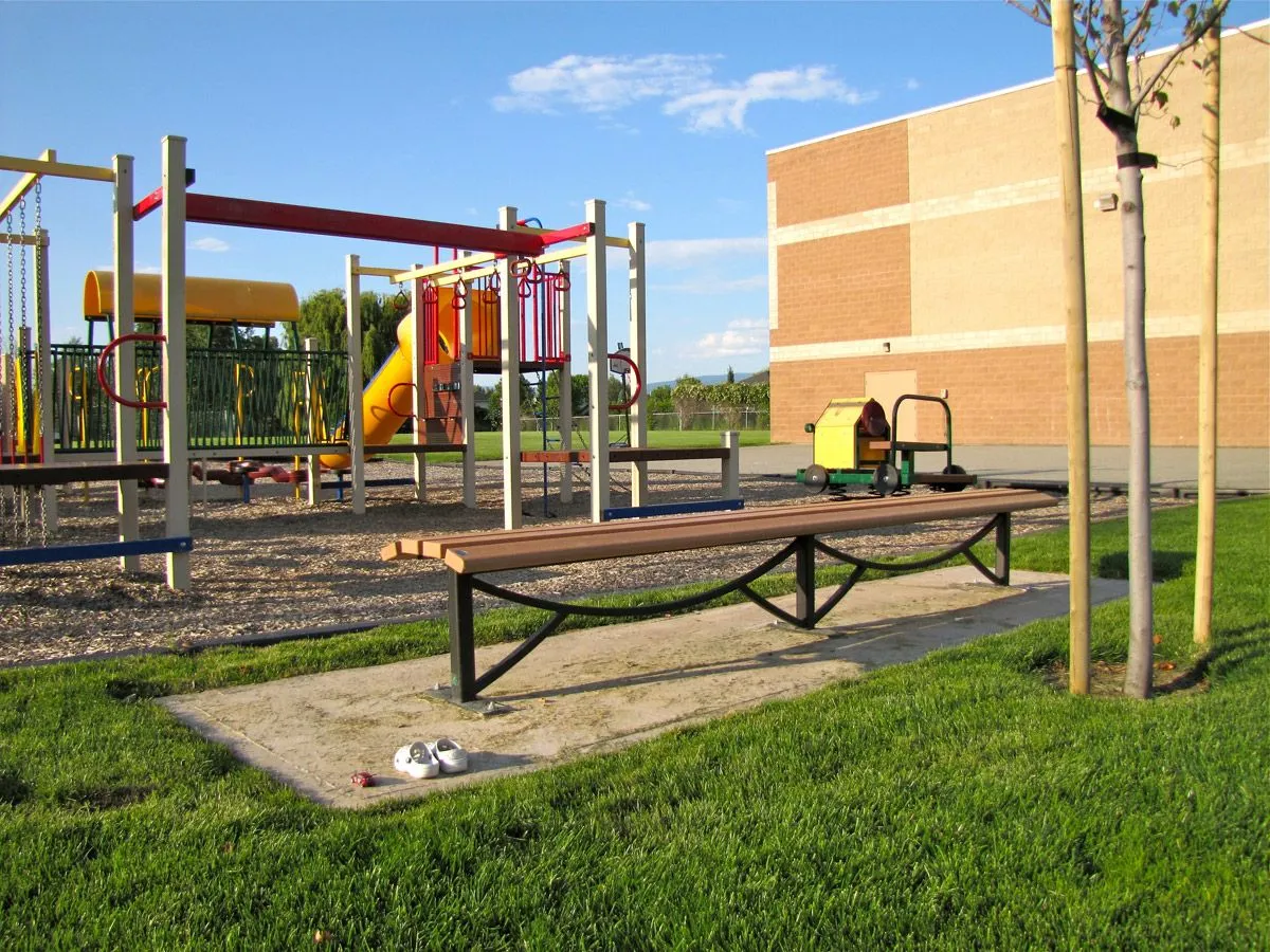 A Thompson bench sits on a concrete pad near a playground and school building. The bench features a curved metal frame and recycled plastic lumber.
