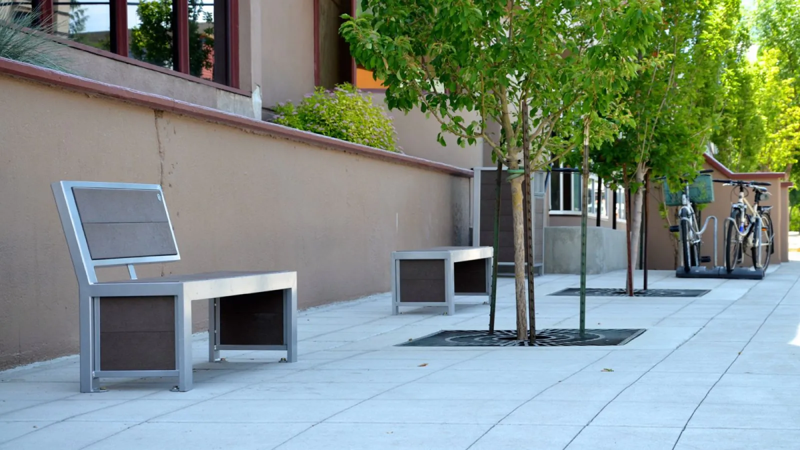 These Urban Form benches provide seating along a paved walkway lined with trees. The benches feature a sleek aluminum frame and recycled plastic lumber.