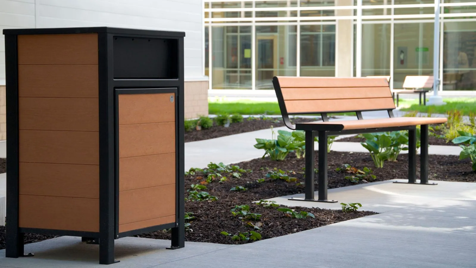 Outdoor setting featuring a waste receptacle and park bench. The furniture is constructed with recycled plastic lumber and powder-coated metal.