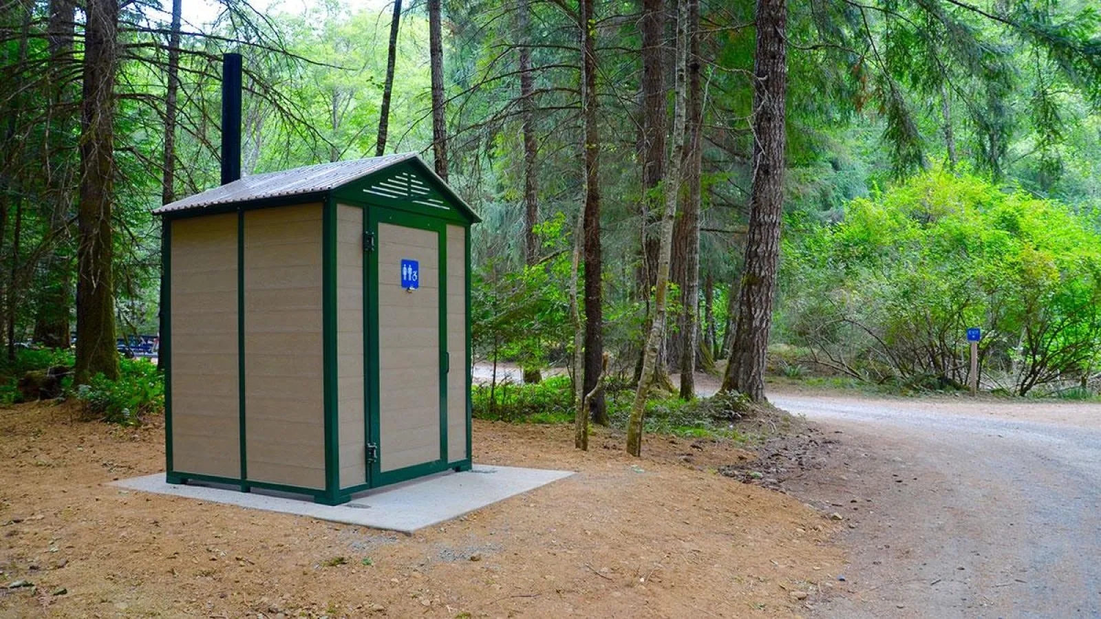 Wheelchair-accessible pit toilet building in a wooded park setting. The structure features tan recycled plastic lumber panels with green trim.