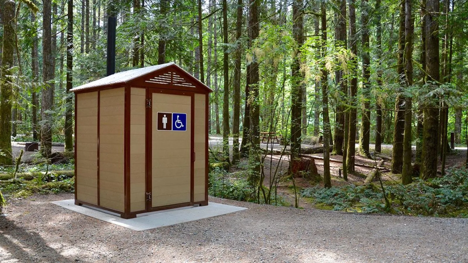 Wheelchair-accessible pit toilet building in a forest setting. The outhouse features a brown frame and tan siding, with signage indicating accessibility.