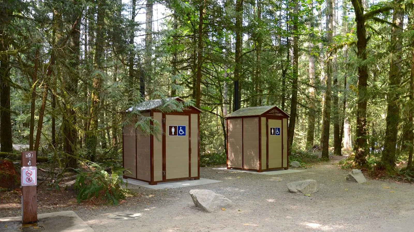 Wheelchair-accessible pit toilets in a forest setting. The structures feature light-colored siding with darker trim.