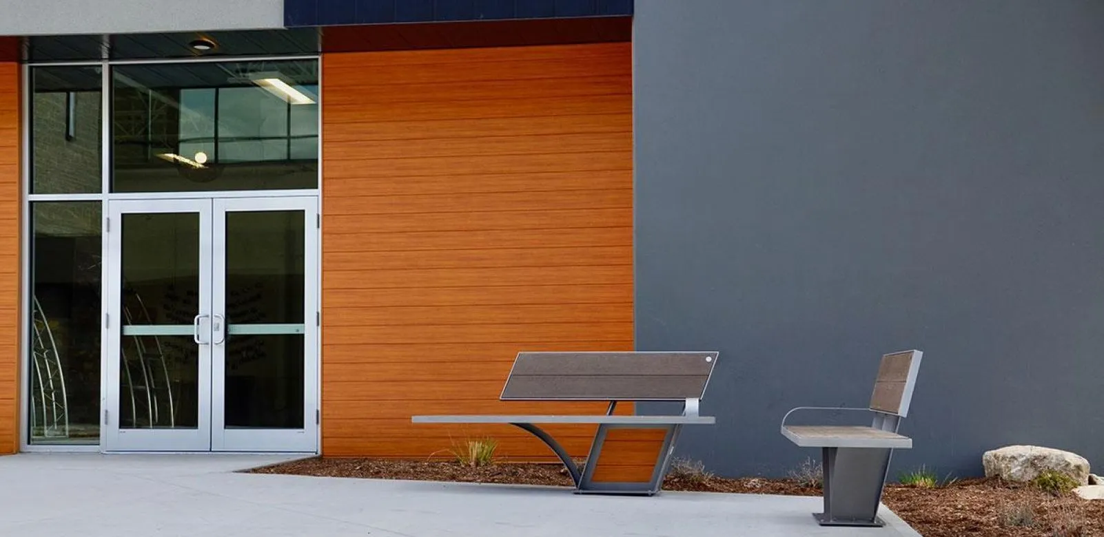 Two benches sit outside a building with orange wood-look siding and a gray wall. The benches feature gray recycled plastic lumber and powder-coated metal frames.