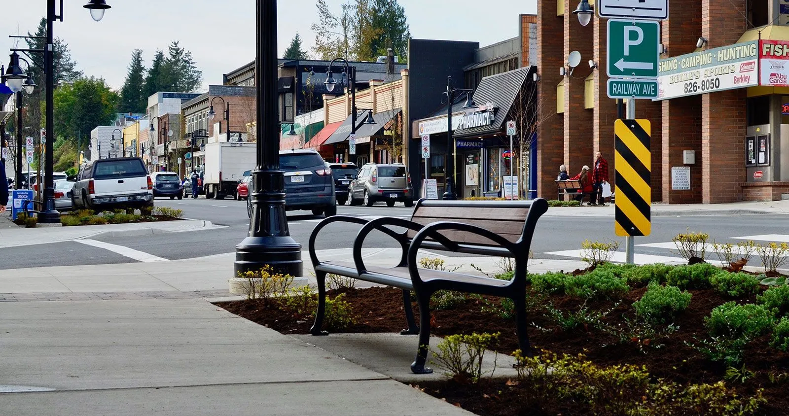 A Mountain Classic Wide Body Bench provides seating along a downtown street. The bench features a black powder-coated frame and wood-look recycled plastic lumber.