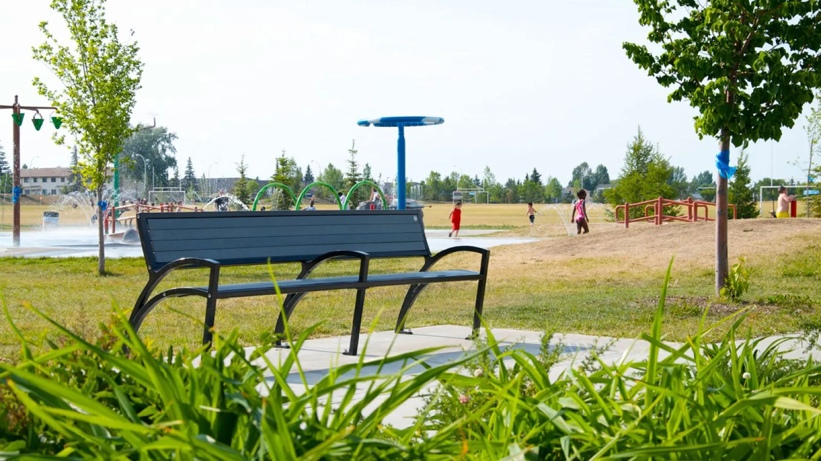 A Modena bench provides seating at the Callingwood Spray Park in Edmonton, Alberta. The bench features recycled plastic lumber and a powder-coated metal frame, offering a comfortable place to rest near the splash pad.