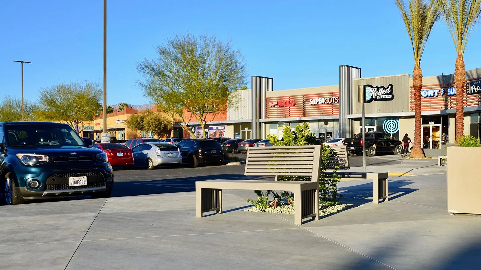 All-aluminum bench with a modern design provides seating in a commercial area. The bench features a slatted back and a neutral finish that complements the surrounding landscape.