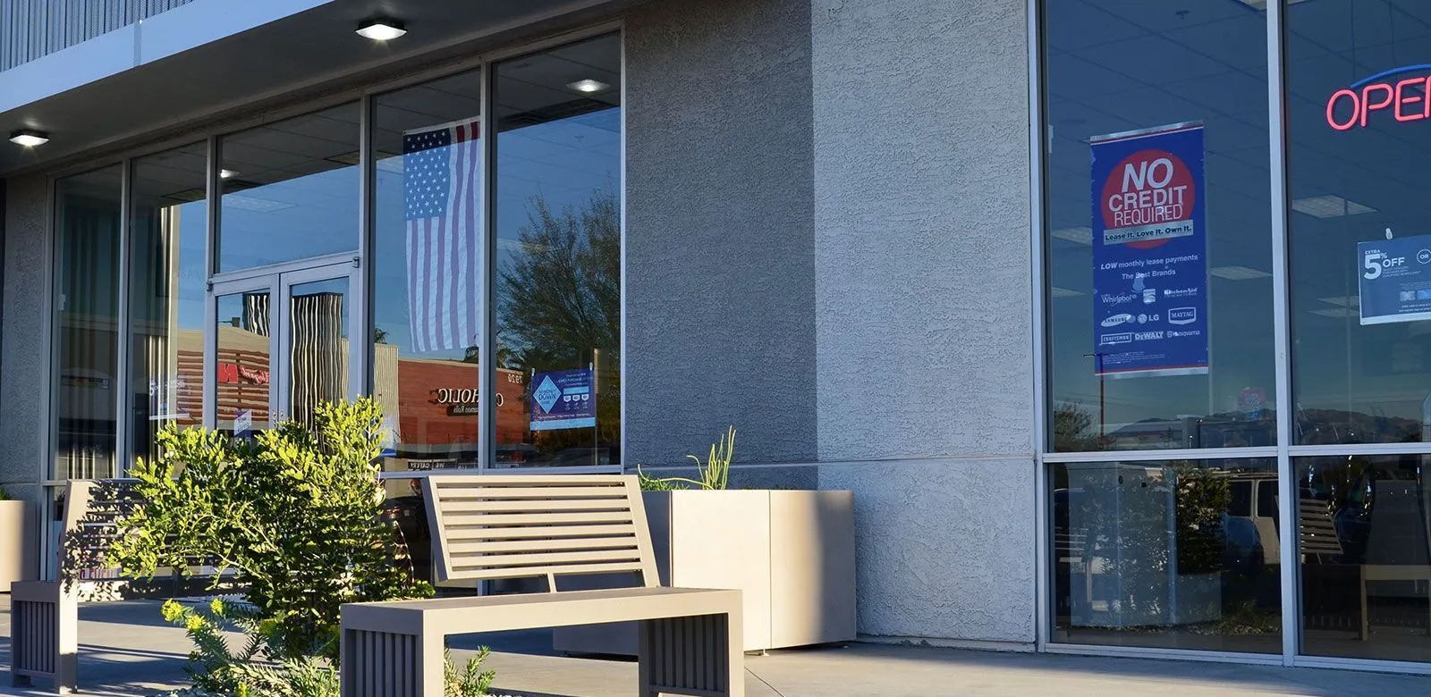 All-aluminum park bench with horizontal slats provides seating outside a commercial building. The bench is powder-coated in a neutral tone and complements the building's modern architecture.