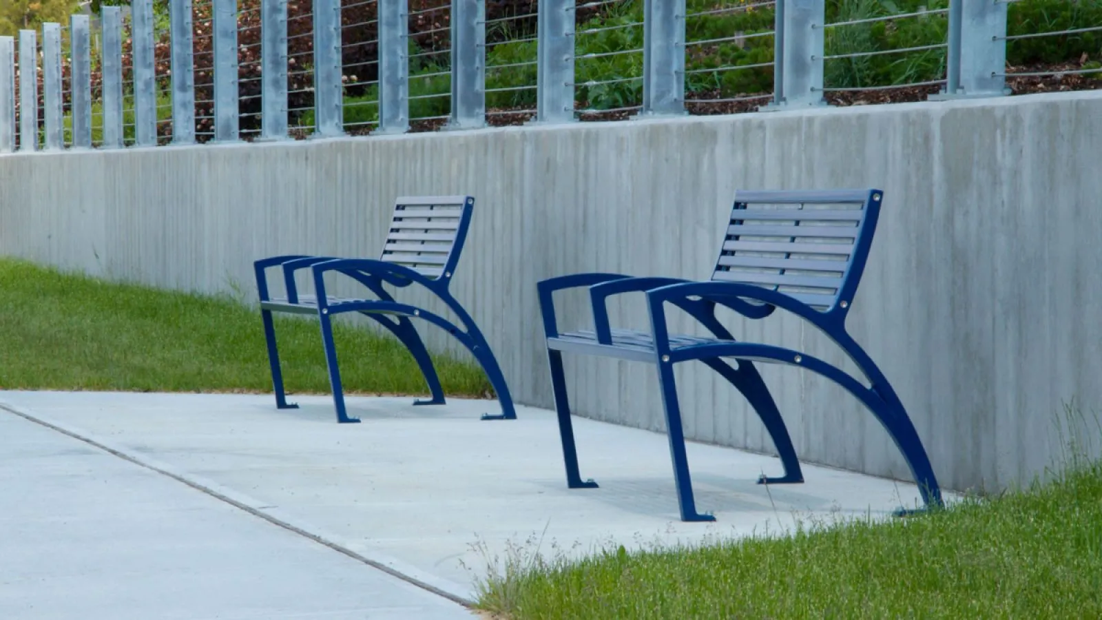 Two metal benches sit along a concrete path next to a low wall with a metal railing. These benches provide seating in a public space.