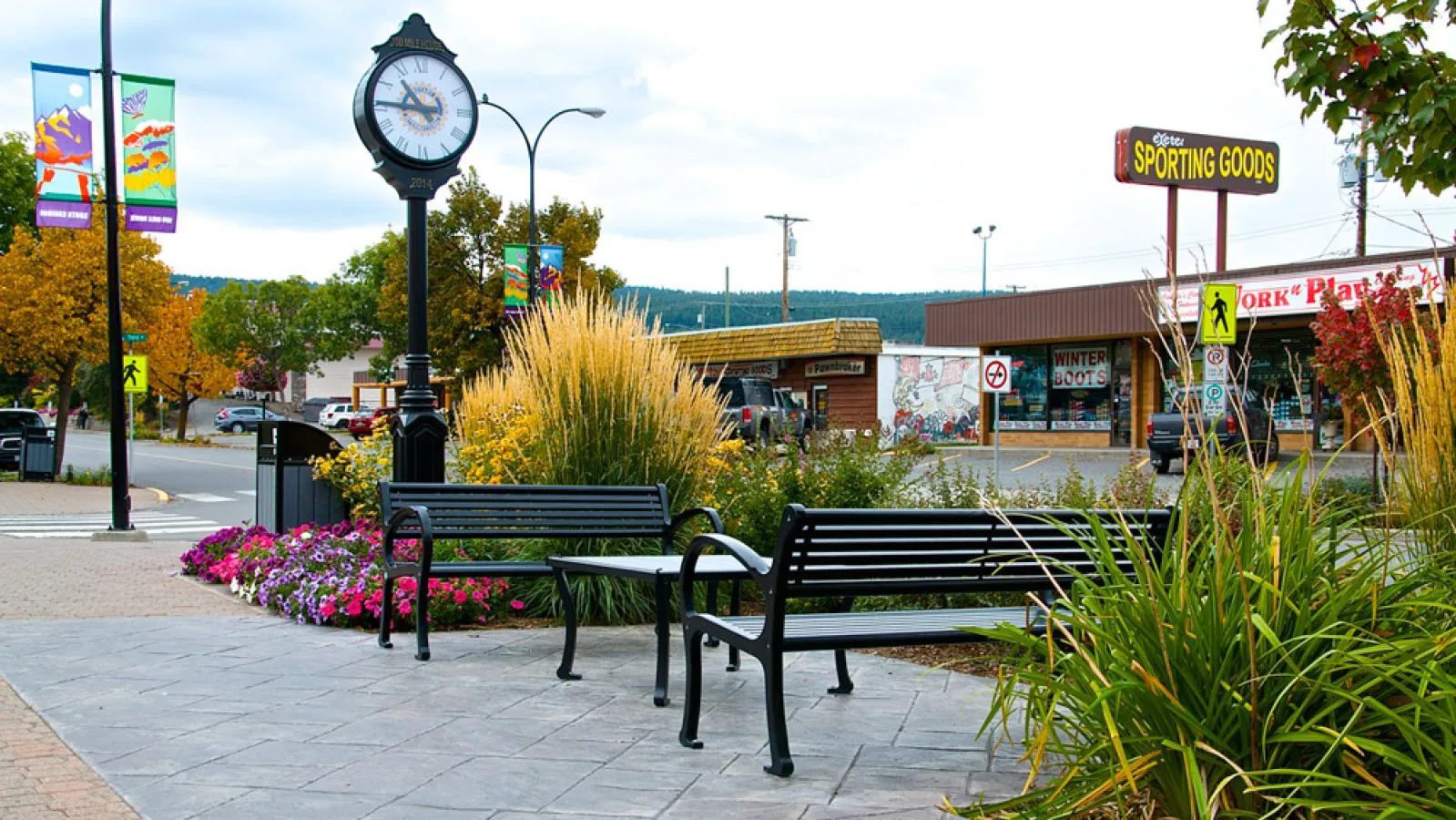 All-metal benches and a coffee table furnish a public space in 100 Mile House, British Columbia. The benches and table provide seating near a flower bed and a clock tower.