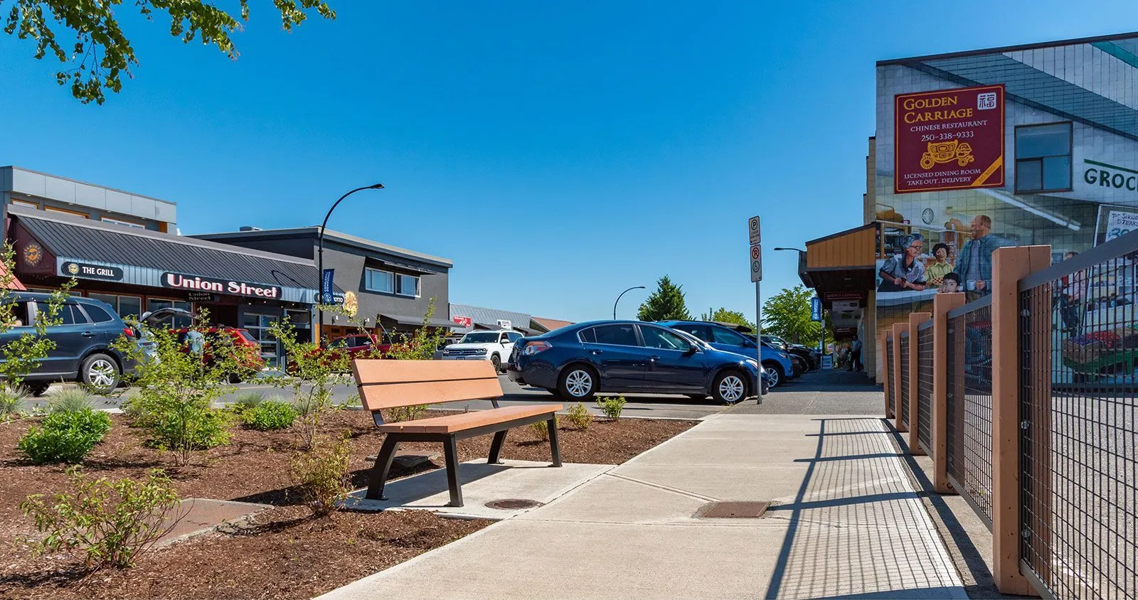An Aylesbury bench provides seating along a sidewalk in Courtenay, British Columbia. The bench features recycled plastic lumber and a powder-coated metal frame.