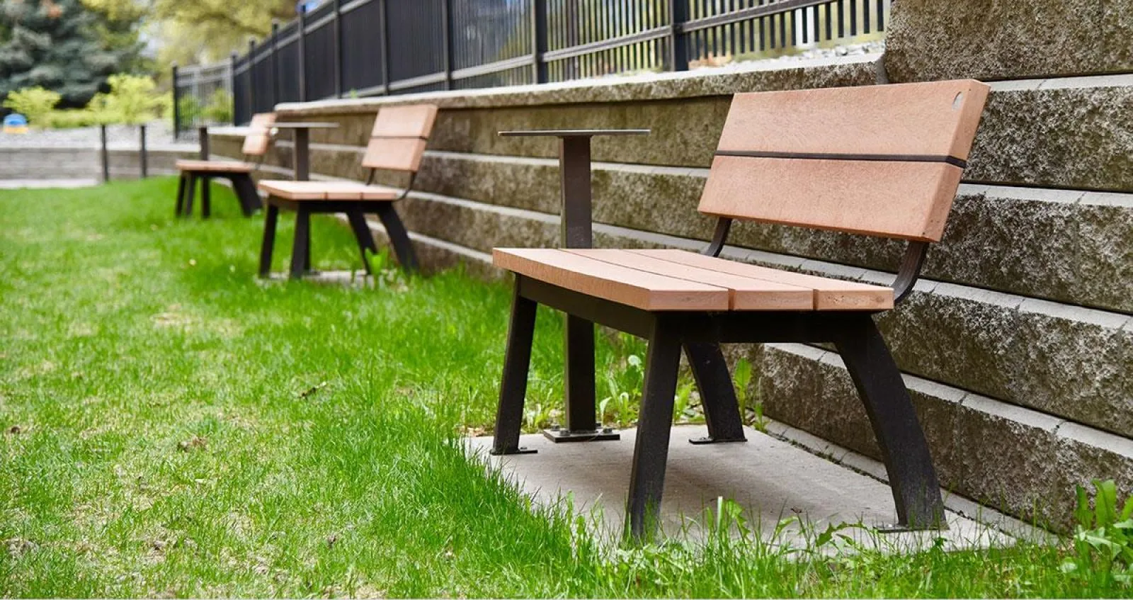 Aylesbury benches with a table are arranged in a row along a low retaining wall in Kelowna, British Columbia. The benches feature recycled plastic lumber seats and backs with a dark powder-coated metal frame.