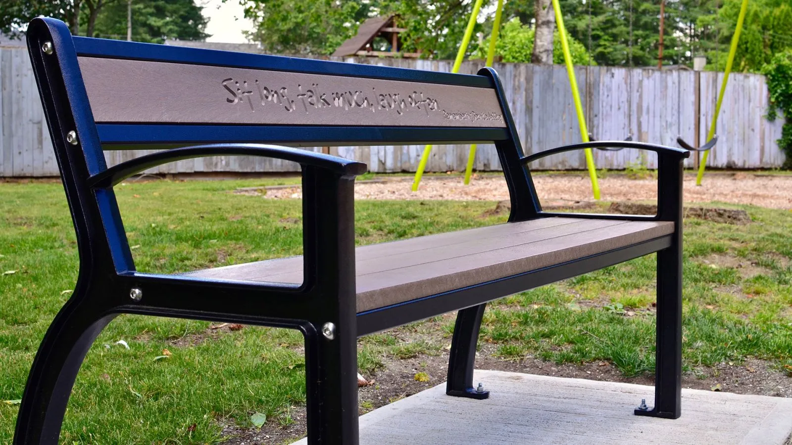 A Beselt bench with a custom engraving sits in a park setting. The bench features recycled plastic lumber and a powder-coated metal frame.