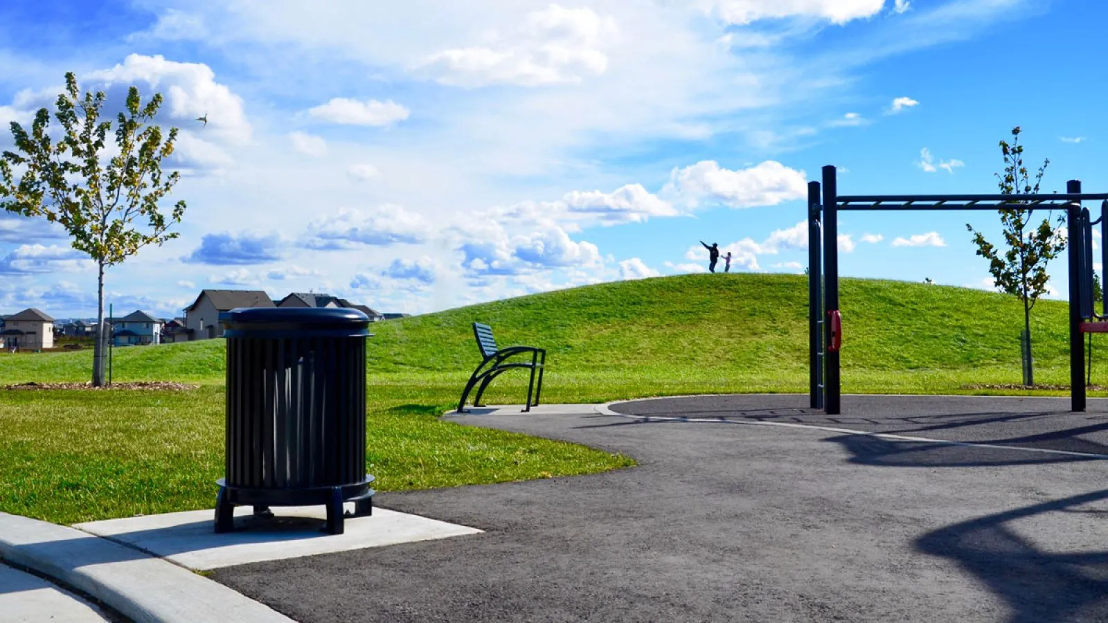 A Beselt Square Top Waste Receptacle sits in a grassy park area with a walking path. The park features a hill with people on top, a bench, and exercise equipment.
