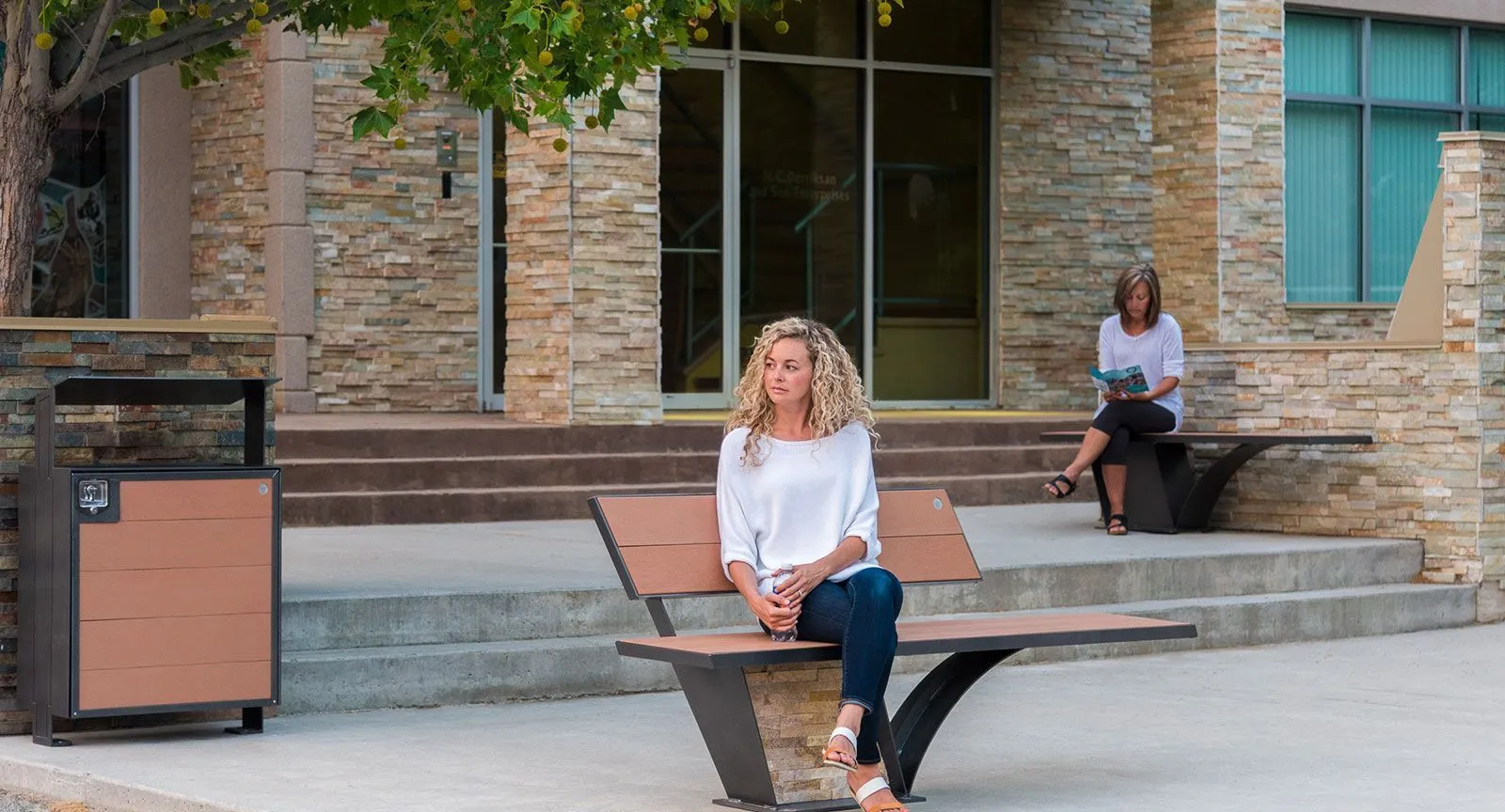 Outdoor benches and a waste receptacle furnish a public space in front of a stone building. The City Scape bench offers a comfortable place to rest, while the waste receptacle helps maintain cleanliness.