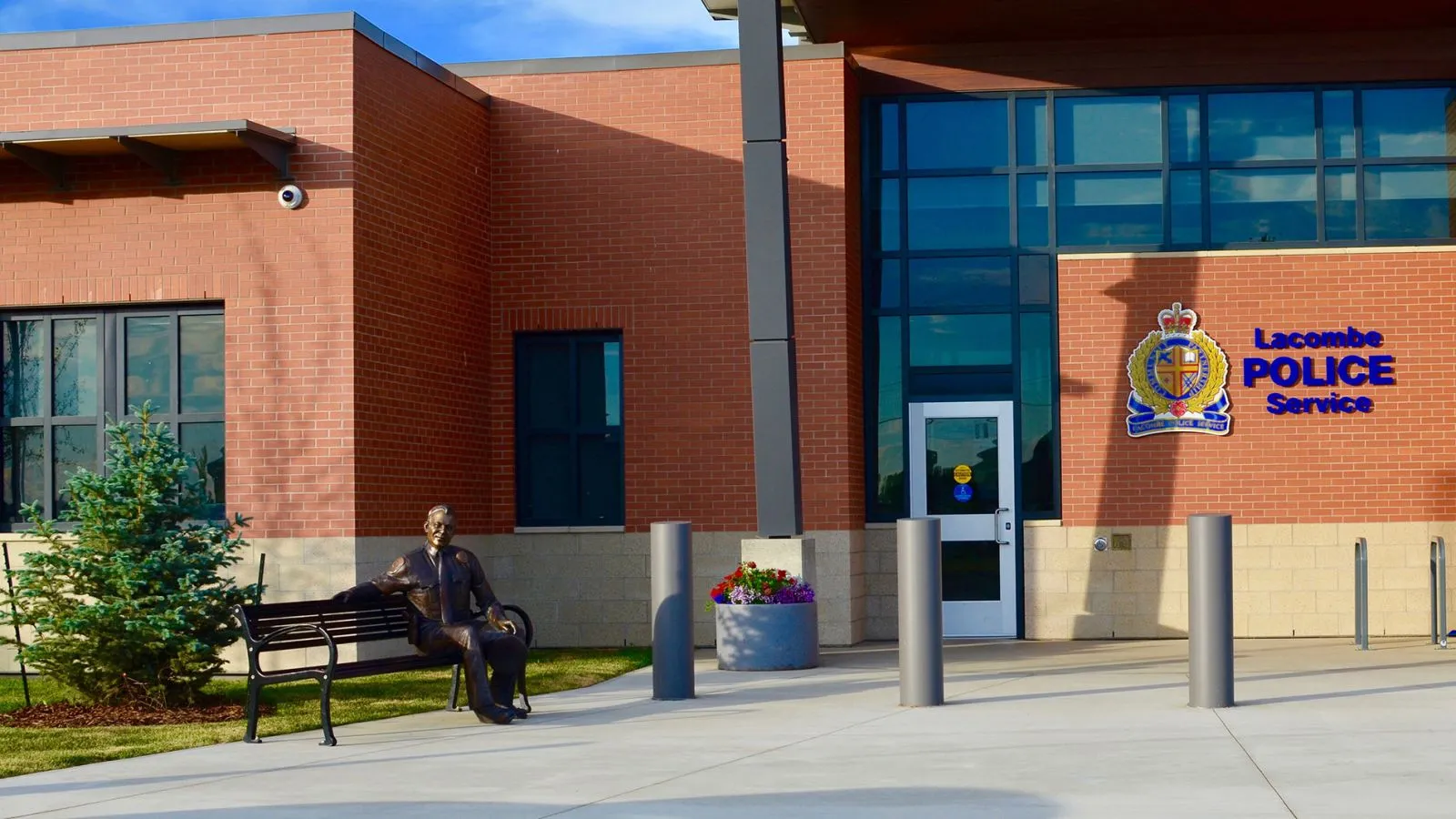 A Mountain Classic bench sits outside the Lacombe Police Service in Alberta, Canada. The bench provides seating near a statue and planter, complementing the building's architecture.