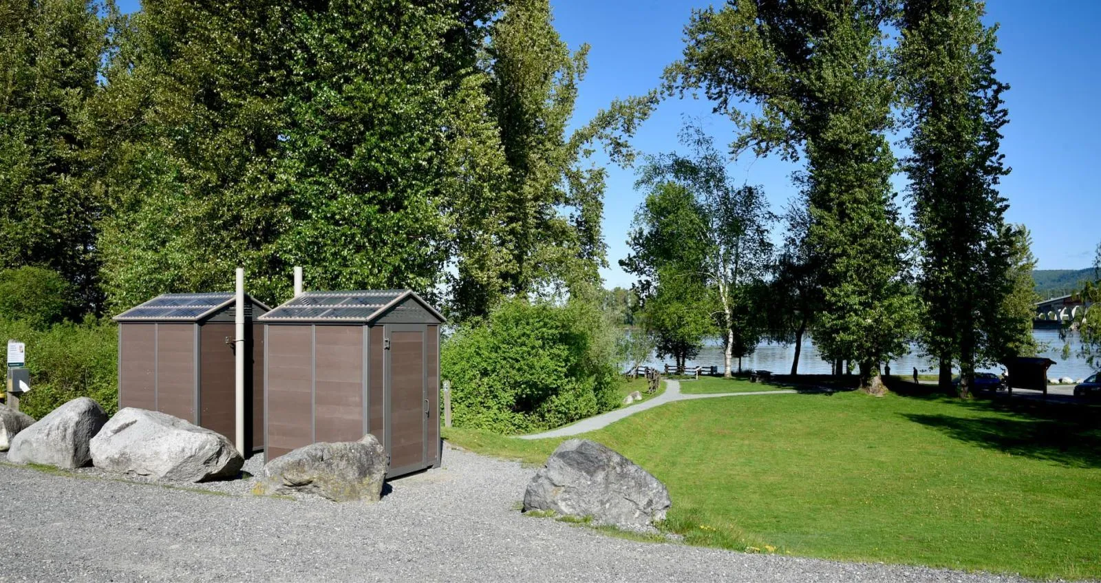 Two jumbo outhouse buildings stand in a park-like setting with a gravel path and view of a lake and bridge in the distance.
