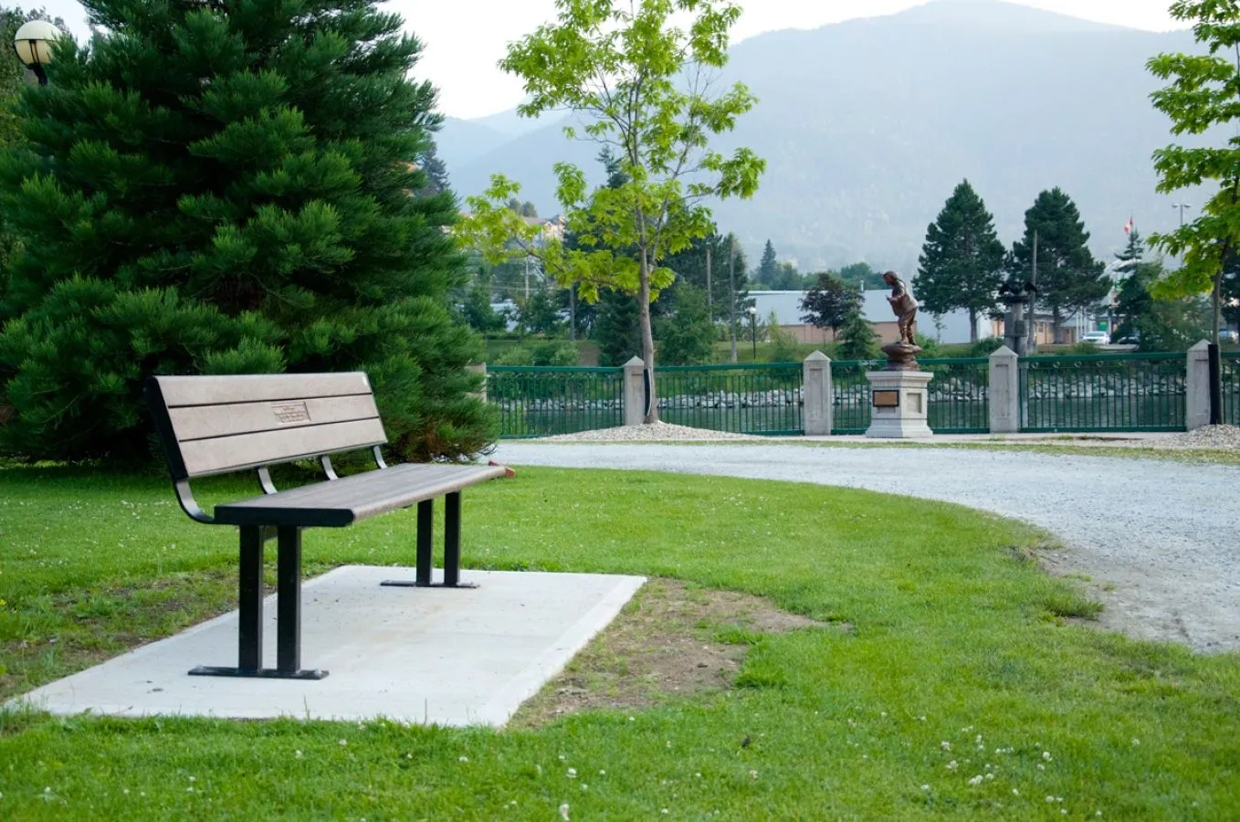 A Larson bench sits in a park setting with a statue and mountains in the background. The bench features recycled plastic lumber and a powder-coated metal frame.