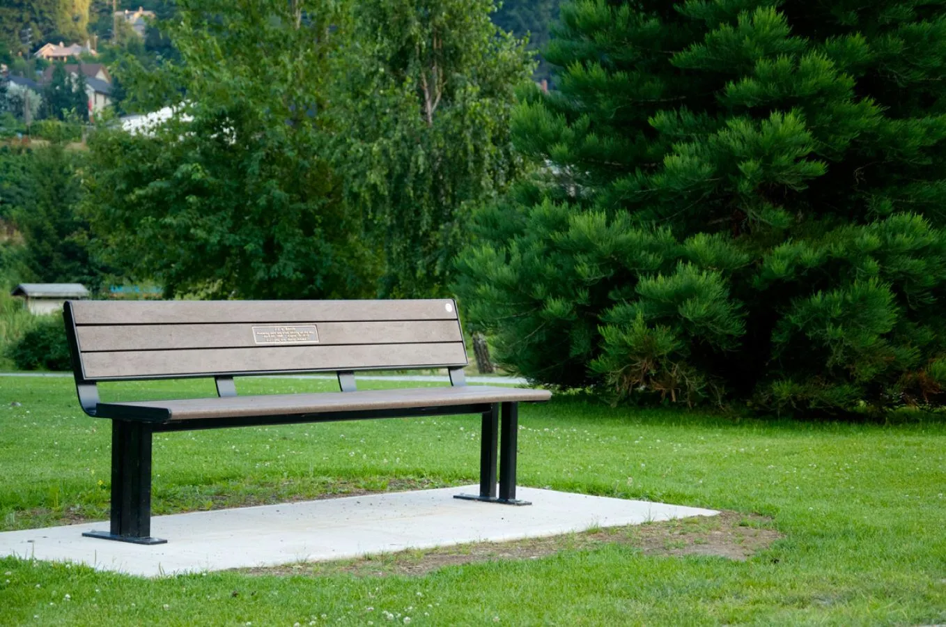 A Larson bench sits on a concrete pad in a grassy park setting. The bench features recycled plastic lumber and a black powder-coated frame.
